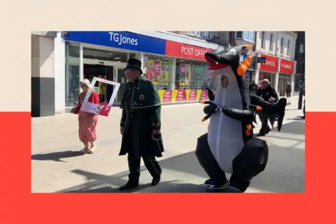 A person dressed in a dragon costume walks through a pedestrianised shopping precinct with TG Jones and Post Office outlets in the background