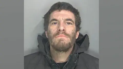 Jason White in a police mug shot. He has curling shortish dark brown hair and a beard. He is wearing a black jacket with its hood pushed down around his neck.