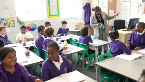 Inside a classroom, where children wearing purple uniform are sitting at their desks listening to a teacher who is addressing the class