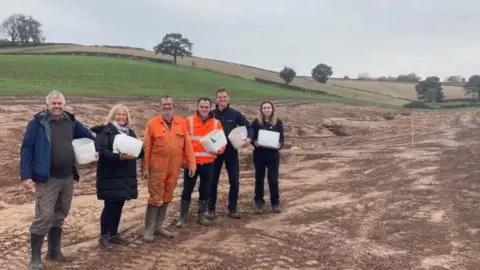 Nottinghamshire County Council Peter Wiles and Amanda Mellor from the flood action group; John Charles-Jones, Landowner, Joshua Wells from the county council and Benjamin Doughty and Amy Facer from the Environment Agency standing in a line in a field where earth has been exposed during work on the small waterway in the background