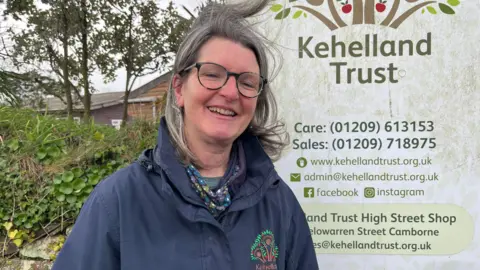 Sally Pyner wearing a blue jacket and stood by a sign for the charity. She has long dark grey hair and glasses. 