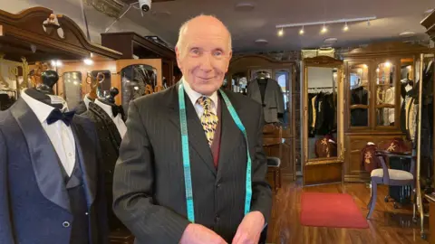 A bald-headed man wearing a pinstriped suit with white shirt and yellow pattern tie and a blue measuring tape around his neck, standing in a tailor's shop with a couple of dummies dressed in blue and black tuxedos to one side.