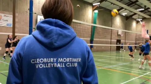 A sports hall with a green floor and people playing volleyball on either side of a high net, with a woman with short brown hair and a blue hoodie in the foreground