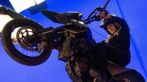 Cameron Diaz does a wheelie on a motorbike against a blue screen