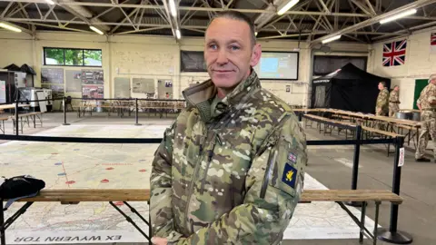 Sgt Tosh Hodgkins stood in the control centre at Okehampton Camp on Dartmoor. There is a big map on the floor behind him and other military personnel in the background. He is wearing camouflage military clothing.