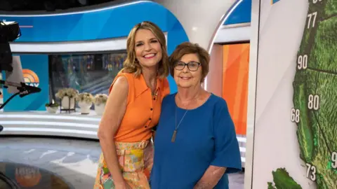 Getty Images Savannah Guthrie smiling widely with a vibrant orange sleeveless blouse and a bright orange and yellow pattern skirt, standing next to her mother in the Today studio. Nancy is smiling, wearing a necklace, and wearing a blue top. 