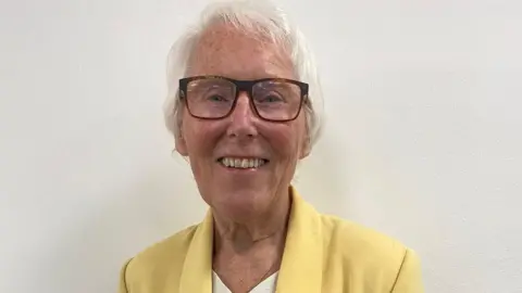 A smiling woman wearing glasses with short, grey hair. She is wearing a cream top underneath a yellow jacket with the top of a yellow rosette showing.