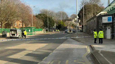 A police road closure - there are two officers stood on the pavement in front of police tape and there is a police car within the cordon further down the road