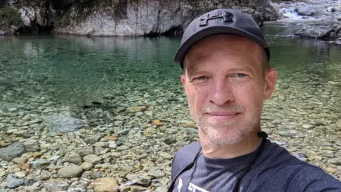 Dr Chris Riley is wearing a blue cap and T-shirt and standing besides a pool of clear water.