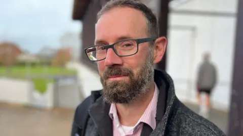 Deputy Aidan Matthews - A man with short black and grey hair. He is wearing black rectangular rimmed spectacles, and is smiling slightly towards the camera. Around his mouth and on his sideburns he has a black and grey beard. He is wearing a grey and white speckled jacket, on top of a red and white buttoned shirt. 