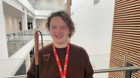 Annabel Amos/BBC A blind student looking directly at the camera. She is stood in front of a white hall wearing a brown jumper with a red lanyard. She has short hair and in her right hand is her cane that is black with a brown handle at the top. 