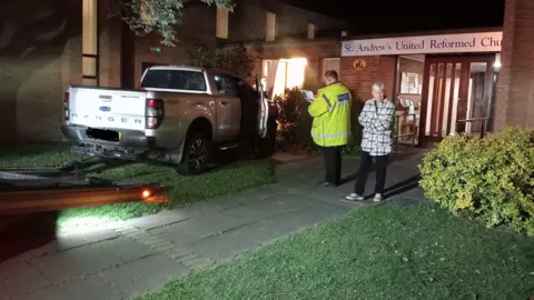 Philip Laws Crash scene- with a police officer in a green police jacket standing next to the vehicle - in front of the church building with a woman in a checked-coat is monitoring the situation from the side of the officer