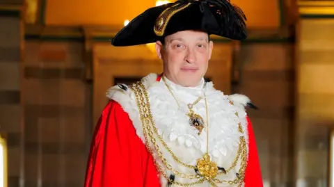 Picture features Cllr Cohen wearing traditional Lord Mayor dress robes. They are red with a white collar and he has a golden chain around his neck. 
He also wears a black hat with a feather. 