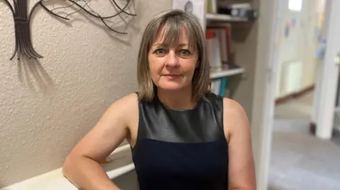A woman looks directly at the camera. She has shoulder length brown hair and is leaning on a shelf. She is wearing a black and silver top.
