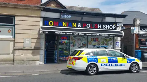 A police car is parked outside a Romanian & Polish Shop. The windows advertise American candy, soft drinks, dairy, snacks, tobacco, and vapes.