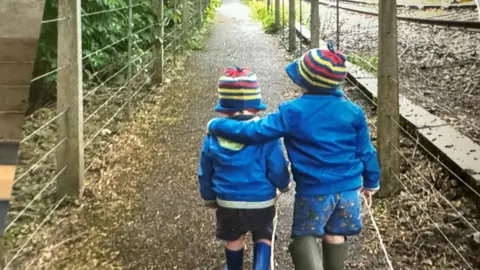 John Hosking Two little boys in blue jackets with knitted hats with red, yellow and white stripes, wearing wellies walking toward the railway station 