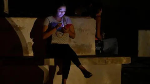 AFP A woman is illuminated by her mobile phone during the third night of a nationwide blackout in Havana October 20, 2024. 