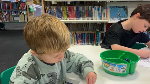 BCP Council Generic image of two young boys in a library colouring in at a table with books behind them in the background.
