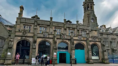 A general view of the entrance to Carlisle railway station. The front door and the window next to it are boarded up. The stone building is reminiscent of a castle with large arched windows and a clock tower with turrets. 