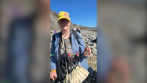 Lyme Regis Museum/Heather Steed Heather is wearing a yellow cap, blue jacket and earthy coloured top, and is wearing a yellow bag slung around her front. She is holding up the fossil to the camera. Her eyes are closed. She is on a rocky beach. 