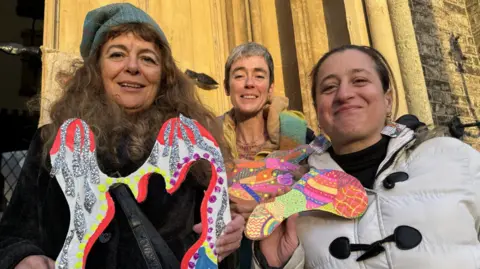 Three women holding up multicoloured cardboard cut-outs and smiling at a camera. On the left a woman wearing a black coat, green hat and with long brown hair. In the middle a woman with short grey hair and a multicoloured scarf and on the right a woman with a white coat. 