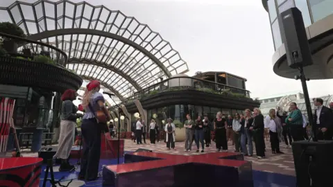 Musicians perform on an outdoor stage in a modern courtyard while a small audience stands watching beneath a large arched glass-and-metal canopy.
