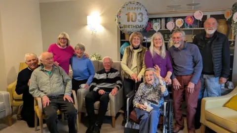 Mavern House Care Home A group of people sat on chairs in a living room, smiling at the camera. They are surrounded by mylar birthday balloons and bunting.
