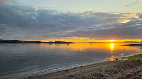 James Boyle The sun sets over a beach and the sea in front of it.