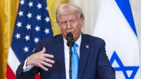 Donald Trump speaking into a mic during a press conference while standing in front of a US and Israel flag