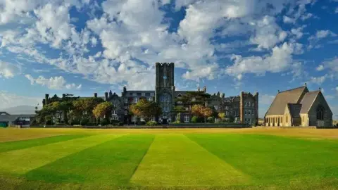 King William's College A neatly mown green lawn in front of a large grey building with a tall clock tower in the middle. A chapel building sits to the right.