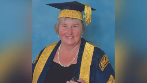 University of Gloucestershire A woman with cheek-length grey hair smiles as she stands for a professional photo at a university event. She is wearing teal and gold robes and a mortarboard, along with pearl jewellery.
