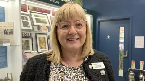 Helen Howell with long blond hair glasses wearing a speckled brown top and brown cardigan standing in front of display cabinets 