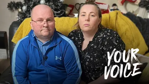 BBC An image of two people sitting on a sofa, with Christmas decorations in the background. On the left is a bald man wearing glasses and a tracksuit jacket; his right leg is a prosthetic decorated with Saltire flags and a crutch leans against the couch next to him. On the right, a woman leaning against him, wearing a patterned shirt, with her hair in a bun. 