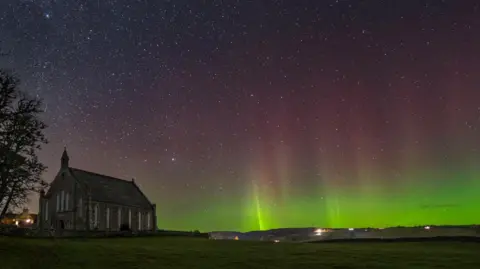 A night-time scene showing a church, which is darkness, and the sky behind the building glowing with a display of the aurora borealis. Stars shine behind the aurora's curtain if green and pink lights.