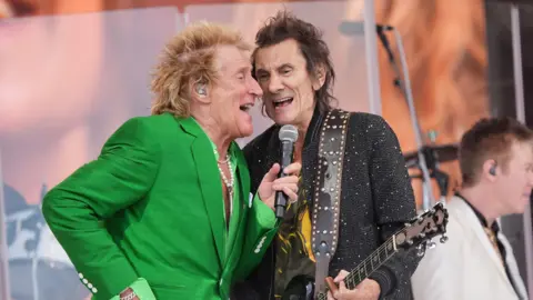PA Media Two men are on stage singing into the same microphone. One of them is wearing a green suit, the other a black jacket.