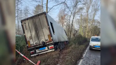 A large lorry, blackened by fire, tilted to one side on a muddy grass verse in a wooded area beside the road. There is a large hole in one side of the lorry. A highways van with an orange light on top is parked by the road next to the vehicle. 