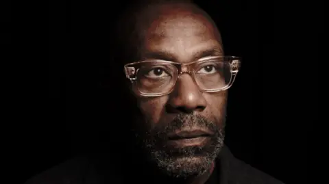 Andy Hollingworth Archive Lenny Henry wears glasses and a beard is looking away from the camera. The background is black.