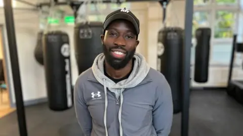 Smiling man in grey jacket and black baseball hat stands in front of punching bags.