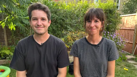 A couple in their thirties sit next to each other in the garden, smiling at the camera. The brunette man wears a dark t-shirt and the woman wears a stripy t-shirt