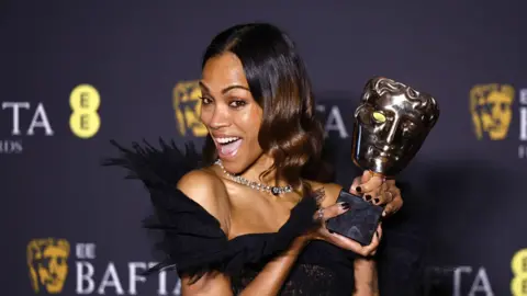 EPA Zoe Saldaña in a black feathery dress holds her Bafta aloft