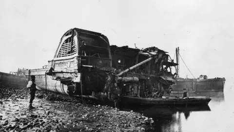 Hulton Archive/Getty Images A black and white image showing parts of a boat run onto a beach next to water. A man stands looking at it.
