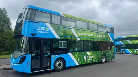 A parked double-decker electric bus with green and blue Translink branding, with the words 'Zero Emissions' and '100% Electric- cleaner air'