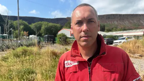 Craig, a man with very short hair is wearing a red jacket with the collar turned up. In the background there's an industrial-looking building and beyond that is a mountainside which has been partially burnt.