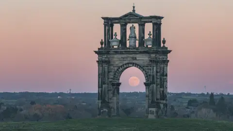The moon is visible through the arch of a monument standing on a field