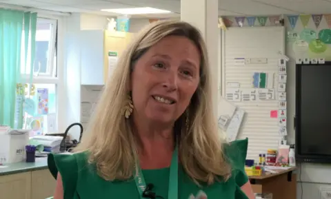 Headteacher Kate Davies is wearing a green sleeveless dress with a lighter green Pines Primary School lanyard round her neck. She is standing in front of a large window with the light streaming through it despite the vertically slatted blind that's pulled partially across it.