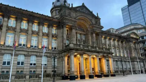 BBC The exterior of the Council House, headquarters of Birmingham City Council. The building has four flagpoles outside it, and comprises of a series of columns and a large archway above the entrance which is lit up by yellow spotlights.
