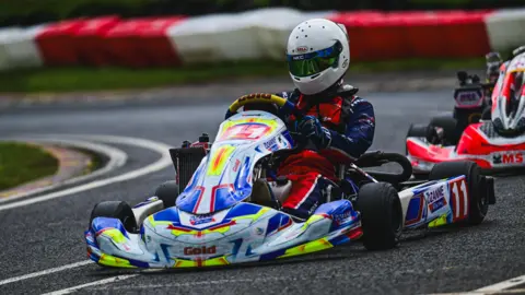 Josh East Photography/Stu Stretton Media A person on a go-kart, which is close to the ground with black wheels and a multi-coloured shell. The person wears a red and blue jumpsuit and a white helmet.