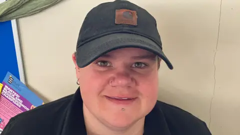 A woman with a nose ring and short hair under a baseball cap is smiling at the camera. She is wearing a dark top.