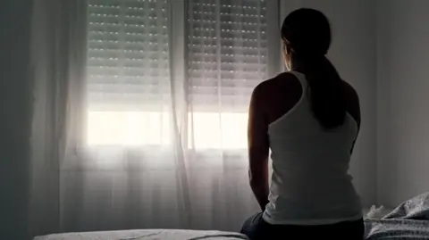 Alvaro Medina Jurado a stock image to represent domestic abuse. A woman with long hair ties back in a low ponytail, wearing a vest top is sat at the edge of a bed in a darkened room. She is facing the window, with a net curtain drawn. Sunlight is visible through the curtain.