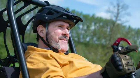 Nick Wilson wears a yellow hoodie and looks into the distance. He is wearing a helmet and gloves holding onto the steering part of the specially designed wheelchair. There is a black overhead structure as part of the chair. He is outside and blue sky and trees are visible. 